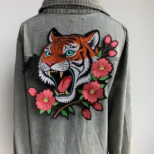 Puede incluir: Chaqueta vaquera gris con un gran bordado de cabeza de tigre con detalles en naranja, blanco y negro, y ojos turquesa. El tigre está rodeado de flores rojas con hojas verdes. La chaqueta tiene cierre de botones y cuello.