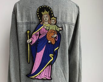 May include: A light-wash denim jacket featuring a large embroidered patch on the back. The patch depicts a religious figure holding a child, with gold, blue, pink, and yellow accents. The jacket has a classic collar and button-down front.