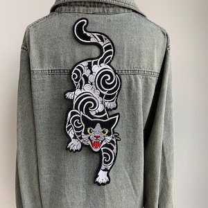 Peut inclure: Une veste en jean gris avec un grand &eacute;cusson brod&eacute; au dos. L'&eacute;cusson repr&eacute;sente un chat noir et blanc avec des motifs tourbillonnants, des yeux jaunes et une gueule ouverte montrant des gencives rouges et des dents blanches pointues.