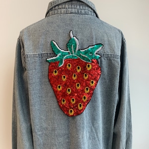Puede incluir: Chaqueta vaquera azul claro con un gran parche de fresa con lentejuelas en la espalda. La fresa es roja con semillas doradas y hojas verdes. La chaqueta tiene un cuello clásico y cierre de botones.