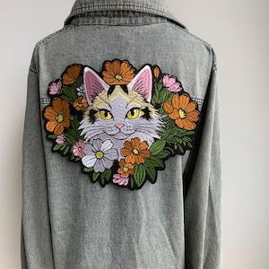 May include: Gray denim jacket featuring a large embroidered patch on the back. The patch depicts a cat's face surrounded by orange, pink, and white flowers. The cat has yellow eyes and a gray and tan fur pattern.