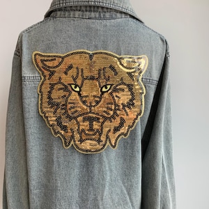 May include: A denim jacket with a gold sequined tiger face patch on the back.