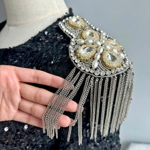 Puede incluir: Charretera decorativa para el hombro con base de lentejuelas negras. La charretera presenta un diseño de cristales transparentes y dorados, perlas y flecos de cadena plateada. El adorno está adherido a una prenda negra.