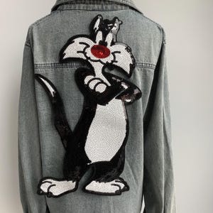 Może przedstawiać: Szara dżinsowa kurtka z dużym czarno-białym haftem z cekinami przedstawiającym kreskówkowego kota na plecach. Kot ma czerwone oczy i białą twarz.