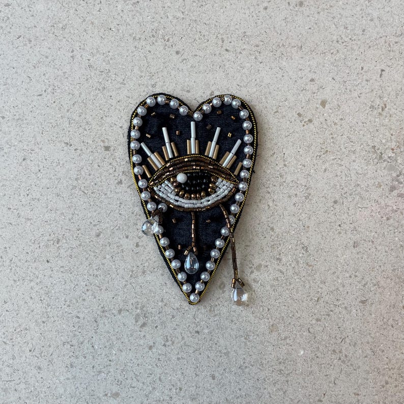 Op de afbeelding: Hartvormige broche met een oogontwerp. De broche is zwart met een kralenrand en een gouden omtrek. Het oog is gedetailleerd met witte, gouden en zwarte kralen. Doorzichtige druppelvormige kralen hangen aan de onderkant.
