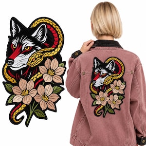 Puede incluir: Un parche bordado que muestra una cabeza de lobo en blanco y negro entrelazada con una serpiente amarilla y flores de color marrón claro con hojas verdes. El parche se muestra en la espalda de una chaqueta vaquera rosa con cuello oscuro.