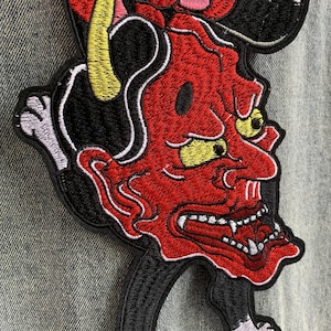 Peut inclure: Un &eacute;cusson brod&eacute; repr&eacute;sentant un d&eacute;mon au visage rouge avec des yeux et des cornes jaunes, des cheveux noirs et des dents blanches ac&eacute;r&eacute;es. Le motif est cousu avec du fil noir, rouge, jaune et blanc sur un tissu en jean textur&eacute;.