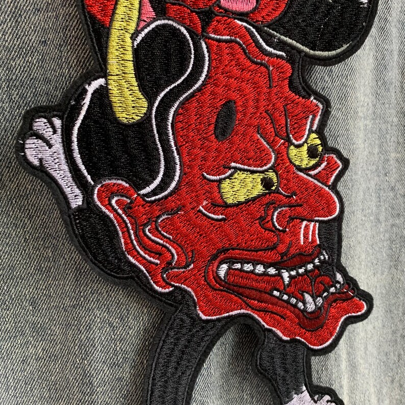 Peut inclure: Un &eacute;cusson brod&eacute; repr&eacute;sentant un d&eacute;mon au visage rouge avec des yeux et des cornes jaunes, des cheveux noirs et des dents blanches ac&eacute;r&eacute;es. Le motif est cousu avec du fil noir, rouge, jaune et blanc sur un tissu en jean textur&eacute;.
