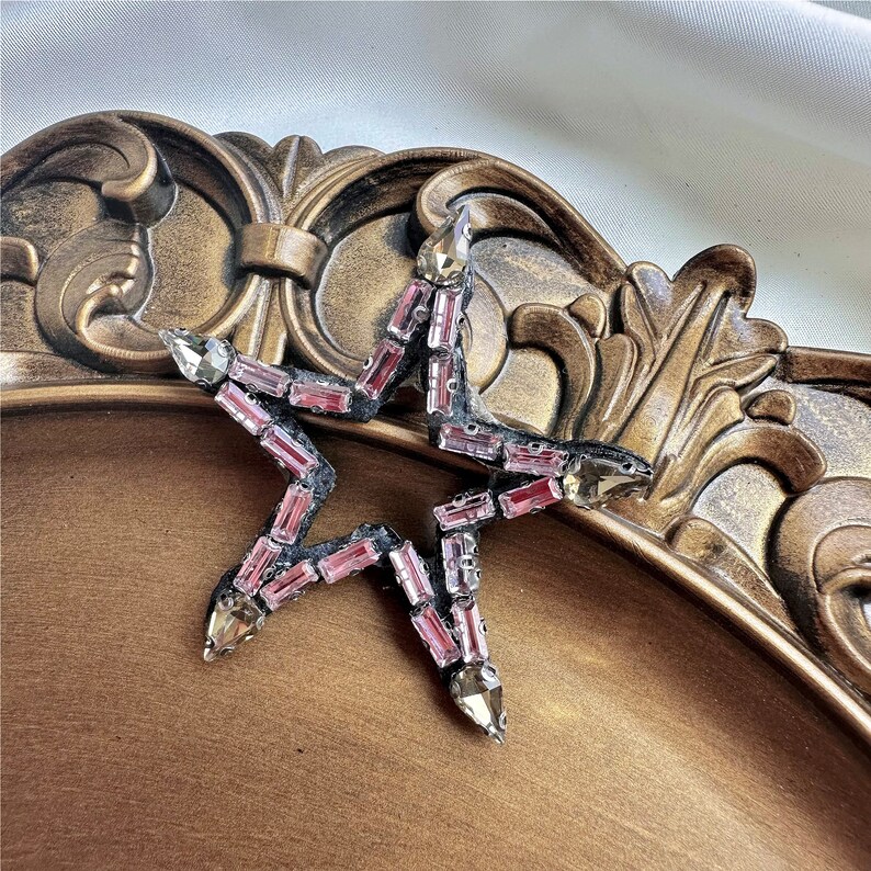 Peut inclure: Une &eacute;toile en strass rose et transparente avec un contour noir. L'&eacute;toile est sur une surface dor&eacute;e.