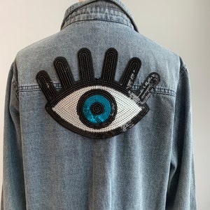 May include: A light blue denim jacket with a large black, white, and blue eye patch on the back. The eye patch is made of sequins and has a blue pupil.