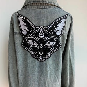 Peut inclure: Veste en jean bleu clair avec un grand écusson brodé représentant un visage de chat dans le dos. L'écusson est noir avec des détails blancs, notamment des étoiles, des lunes et des motifs complexes. La veste a un col classique et une fermeture boutonnée.