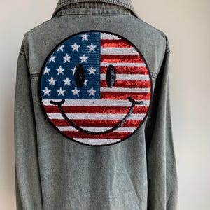 May include: A denim jacket with a large sequined smiley face patch in the shape of the American flag. The patch is red, white, and blue.