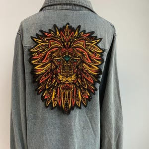 Puede incluir: Una chaqueta vaquera con un gran parche de león colorido en la espalda. El león está representado en un estilo psicodélico con detalles intrincados y un tercer ojo.