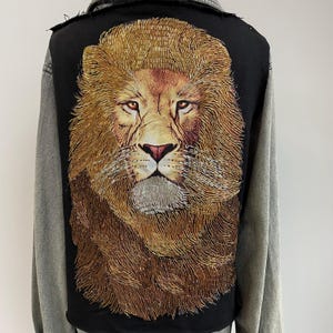 Puede incluir: Chaqueta vaquera negra con una gran cabeza de león bordada en la espalda. La melena del león es de color marrón dorado, y la chaqueta tiene mangas grises y un cuello desgastado.