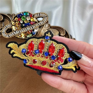 Może przedstawiać: Złota korona wyszywana czerwonymi i niebieskimi klejnotami. Korona jest otoczona czarnym tłem z tkaniny.