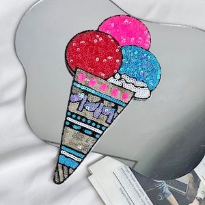 May include: A colorful ice cream cone patch with sequins. The cone is blue, white, and black with a pink, red, and blue ice cream scoop on top.