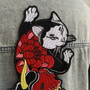 Peut inclure: Un &eacute;cusson brod&eacute; repr&eacute;sentant un chat noir et blanc &agrave; l'expression s&eacute;v&egrave;re, orn&eacute; d'un grand motif floral rouge sur le corps et d'une queue jaune, cousu sur un tissu en jean bleu clair.