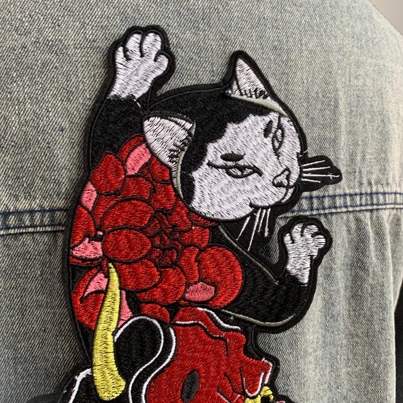 Peut inclure: Un &eacute;cusson brod&eacute; repr&eacute;sentant un chat noir et blanc &agrave; l'expression s&eacute;v&egrave;re, orn&eacute; d'un grand motif floral rouge sur le corps et d'une queue jaune, cousu sur un tissu en jean bleu clair.