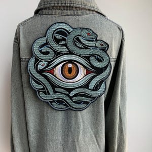 May include: A denim jacket with a large embroidered patch on the back. The patch features a detailed design of a stylized eye with a brown iris, surrounded by intertwined snakes in shades of green and black.