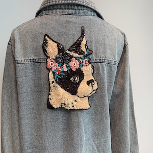 May include: A denim jacket with a sequined dog patch on the back. The dog is wearing a flower crown and has a black nose and eyes.