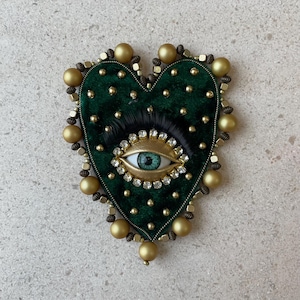Op de afbeelding: Een hartvormige broche met een donkergroene fluwelen basis, versierd met gouden kralen en een centraal oogontwerp. Het oog heeft een lichtblauwe iris, omgeven door goud en heldere kristallen, en zwarte wimpers. De broche is ongeveer 7.6 cm breed.