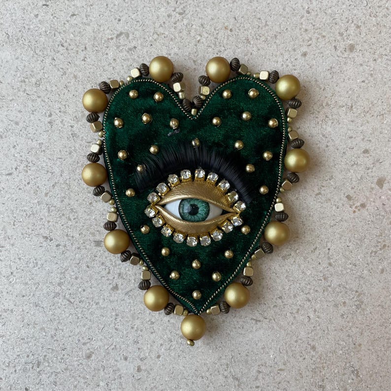 Op de afbeelding: Een hartvormige broche met een donkergroene fluwelen basis, versierd met gouden kralen en een centraal oogontwerp. Het oog heeft een lichtblauwe iris, omgeven door goud en heldere kristallen, en zwarte wimpers. De broche is ongeveer 7.6 cm breed.