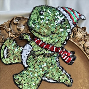 May include: A green sequined dinosaur wearing a red and white striped scarf and a red, white, and green striped hat. The dinosaur is standing on a brown surface.