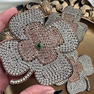 Könnte beinhalten: Ein großer, vierblättriger Kleeblatt-Patch aus Strasssteinen. Das Kleeblatt besteht aus klaren und rosafarbenen Strasssteinen, mit einem einzigen grünen Strassstein in der Mitte.