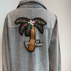 Könnte beinhalten: Eine blaue Jeansjacke mit einem Pailletten-Palmenbaum-Patch, auf dem "Paradise Island" steht.