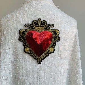 Può includere: Una giacca bianca di paillettes con una toppa ricamata a forma di cuore dorata e rossa. Il cuore è circondato da una cornice dorata decorativa.