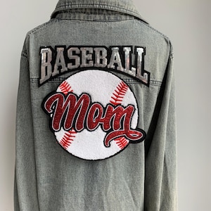May include: A grey denim jacket with a large baseball patch on the back. The patch features the word "BASEBALL" in silver sequins above a baseball with the word "Mom" in red script.