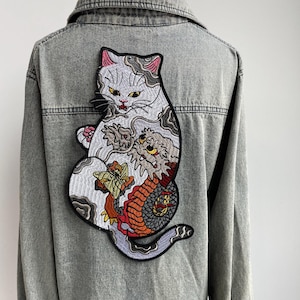May include: Gray denim jacket featuring a large embroidered patch of a cat with a dragon design. The patch includes shades of gray, pink, red, orange, and yellow. The jacket has a classic collar and button-up front.