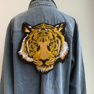 May include: A denim jacket with a large embroidered tiger head patch on the back. The tiger has a brown and yellow striped pattern with white accents and green eyes.