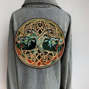 May include: A denim jacket with a large, circular embroidered patch featuring a detailed tree design. The patch has a dark green background with a gold, teal, and orange tree of life design. The jacket is a light blue wash.