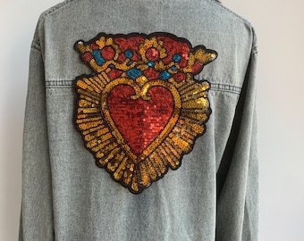 Pode incluir: Casaco de ganga de lavagem clara com um grande patch em forma de coração com lantejoulas nas costas. O patch é vermelho, dourado, azul e laranja, com um design de coração. O casaco tem uma gola clássica e fecho de botões.
