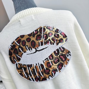 Può includere: Maglione bianco con un grande design a forma di labbra con paillettes e stampa leopardata sul retro.