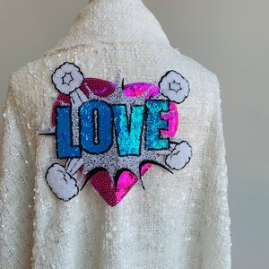 Peut inclure: Veste blanche à paillettes avec un cœur en paillettes rose et bleu et le mot "LOVE" dans un style bande dessinée.