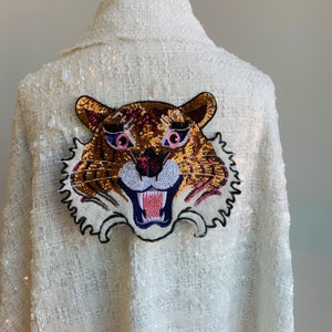 Op de afbeelding: Witte sequinge jas met een grote geborduurde tijgergezichtspatch in goud, roze en paars.