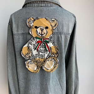 May include: A light blue denim jacket with a brown teddy bear embroidered on the back. The teddy bear is wearing a red and green bow and has the words "If For Fun" embroidered on its chest.