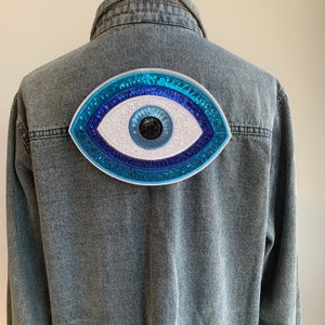 May include: A blue denim jacket with a large, white and blue evil eye patch sewn on the back. The evil eye is made of sequins and has a black pupil.