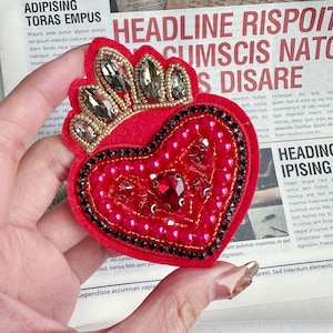 Puede incluir: Un corazón de fieltro rojo con una corona dorada y cuentas rosas y negras. El corazón está decorado con un gran cristal rojo en el centro.