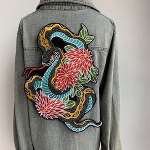 Op de afbeelding: Lichtgrijze spijkerjas met een groot geborduurd embleem. Het embleem toont een zwarte slang met gele en blauwgroene accenten, een rode tong en rode en witte bloemen.