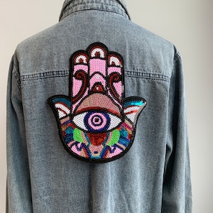 以下が含まれることがあります： 背面にカラフルなスパンコールで飾られた大きなハmsaハンドパッチが付いたデニムジャケット。ハmsaハンドはピンク、ブルー、グリーン、レッドで、中央に大きなブルーアイがあります。