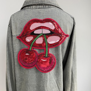 May include: A gray denim jacket with a large sequined patch of red lips and cherries on the back. The lips are pink and red, and the cherries are a deep red color. The patch is centered on the back of the jacket.