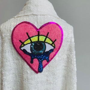 Könnte beinhalten: Weiße Jacke mit Pailletten und einem rosafarbenen herzförmigen Patch mit einem blauen Auge mit gelben Wimpern und blauen Pailletten, die aus dem Auge tropfen.