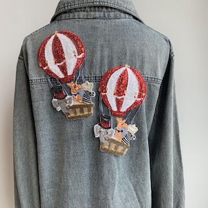 May include: A denim jacket with two embroidered patches of hot air balloons with animals in baskets. The balloons are red and white with sequins. The animals are a giraffe, zebra, elephant, and a bear.