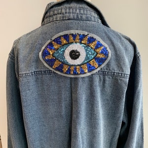 May include: A blue denim jacket with a large, colourful eye patch on the back. The eye patch is made of sequins and features a blue eye with a white pupil and gold accents.