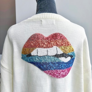 Puede incluir: Suéter de punto blanco con un gran diseño de labios de lentejuelas arcoíris en la espalda.