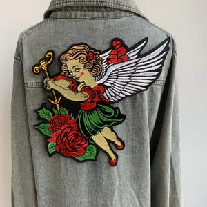 May include: A gray denim jacket with a large embroidered patch on the back. The patch features a vintage-style angel with red, green, and gold details, holding a golden instrument, surrounded by red roses and white wings.