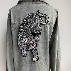 May include: A gray denim jacket featuring a large embroidered tiger patch on the back. The tiger is depicted in shades of gray, black, white, and yellow, with its mouth open. The jacket has a classic collar and button-down front.